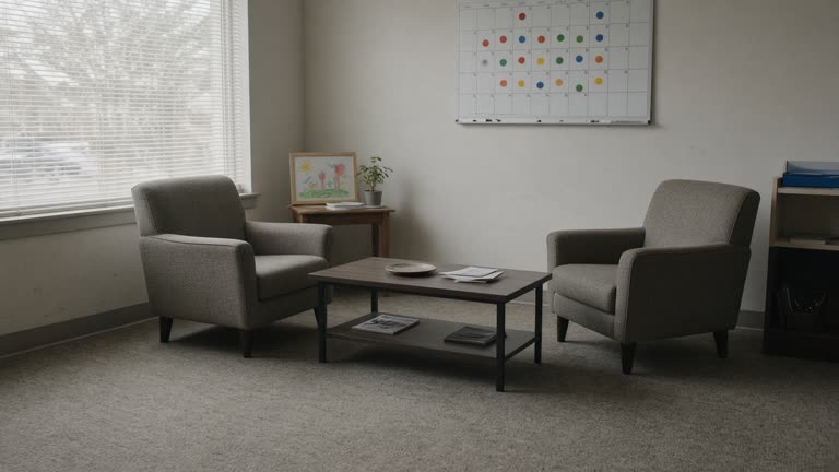 Community counseling room with a wall calendar marked in colored stickers, two chairs facing a low table, soft daylight through blinds, children's drawing on side shelf, no logos or readable text