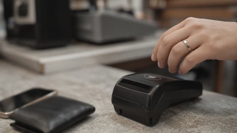 Close view of a plain contactless payment ring on a finger above a café counter terminal, soft indoor daylight, blurred wallet and phone nearby, no logos or readable text on devices