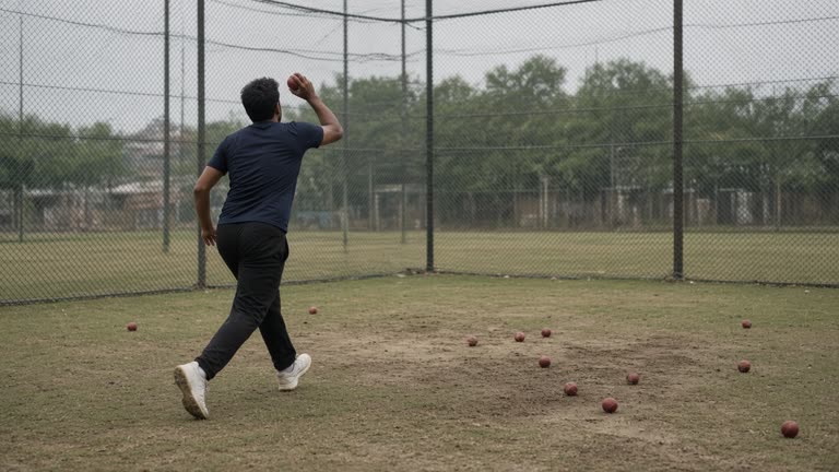 Cricket practice nets with red balls scattered on worn grass, a pace bowler mid-run-up seen from behind without a visible face, overcast humid light and a chain-link fence, no logos or readable text