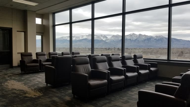 Airport lounge interior with empty leather chairs facing floor-to-ceiling windows showing distant Rocky Mountains under overcast light, carpet wear near the entrance, no logos or readable text