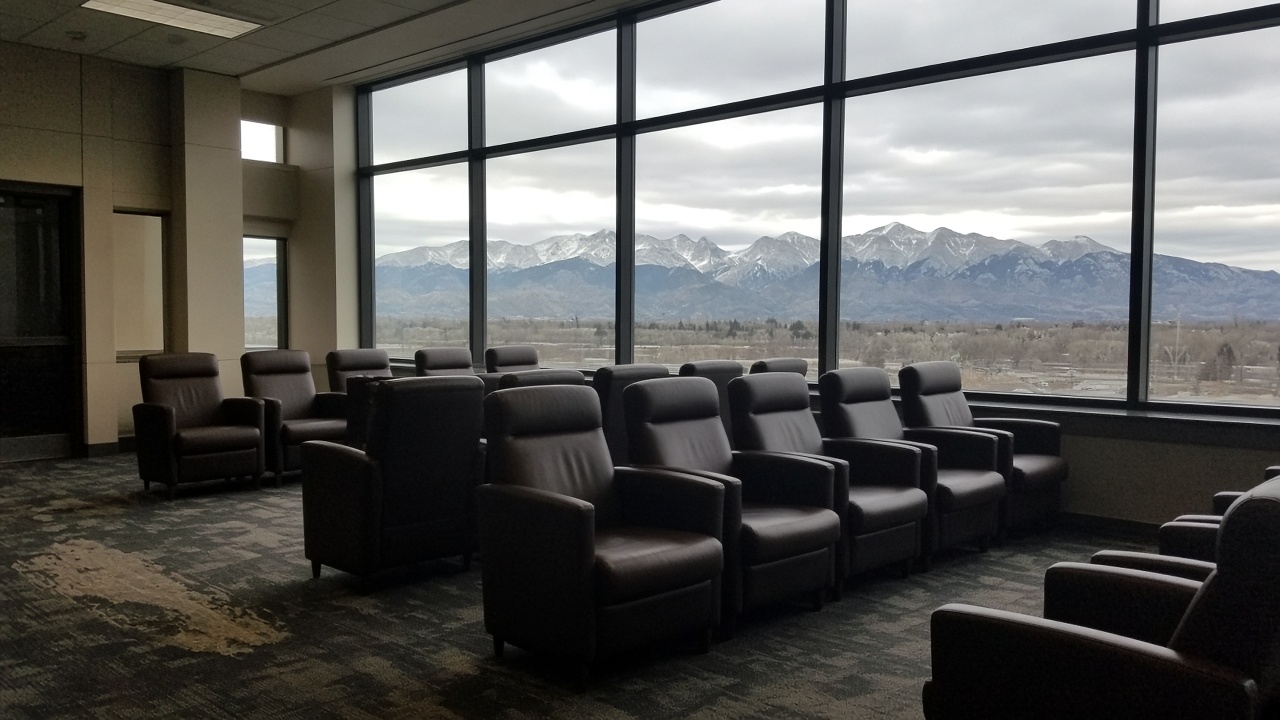 Airport lounge interior with empty leather chairs facing floor-to-ceiling windows showing distant Rocky Mountains under overcast light, carpet wear near the entrance, no logos or readable text