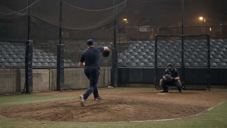 Professional baseball relievers warming up in a bullpen mound area under evening stadium floodlights, scuffed dirt and catching nets, distant empty seats, no logos or readable text