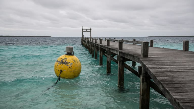 A yellow oceanographic buoy moored near a remote atoll research pier with turquoise water and overcast sky, gentle swell and weathered wood decking, no logos or readable text