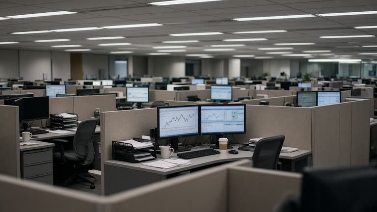Bank operations floor with cubicle dividers and dual monitors showing blurred charts, cool fluorescent light, coffee cups and paper trays, no logos or readable text