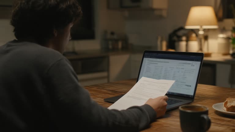 A person reviewing a credit card bill on a laptop at a kitchen table, screen content blurred, warm evening lamp light, no logos or readable text