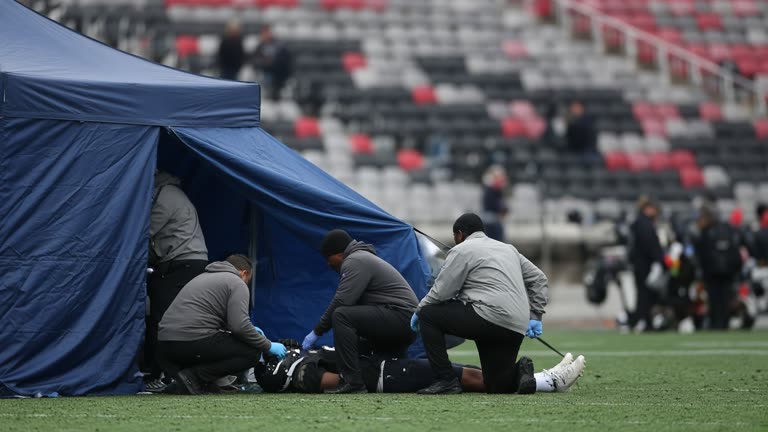 Professional football sideline with blue medical tent and trainers in neutral colors, stadium seats blurred in background, overcast daylight, no team logos or readable jersey numbers