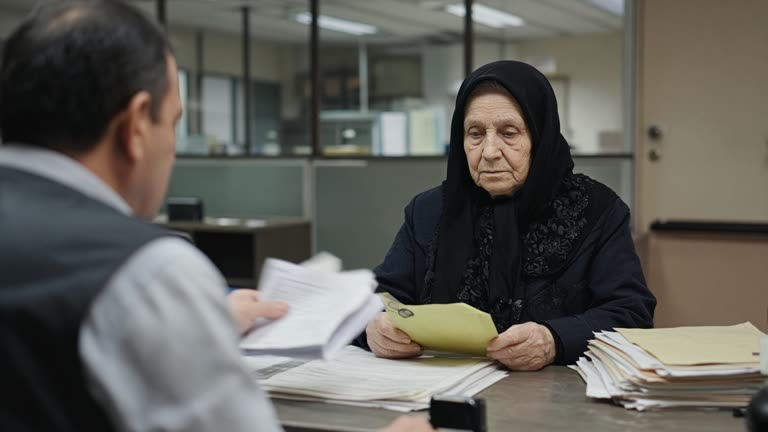 Elderly widow at a municipal tax assistance desk with stacked paper forms and a pension envelope, soft fluorescent light, counselor from behind, no logos or readable text.