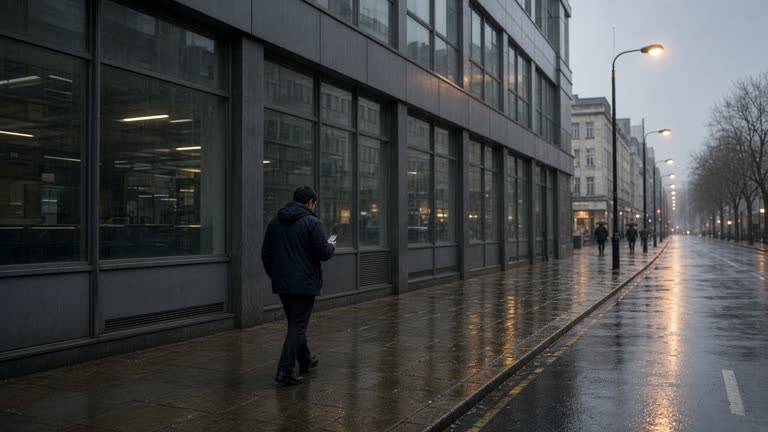 Bristol city centre office block facade in grey rain, commuter reading a phone on wet pavement, blurred handset screen, sodium street lamps reflecting on tarmac, no logos or readable text