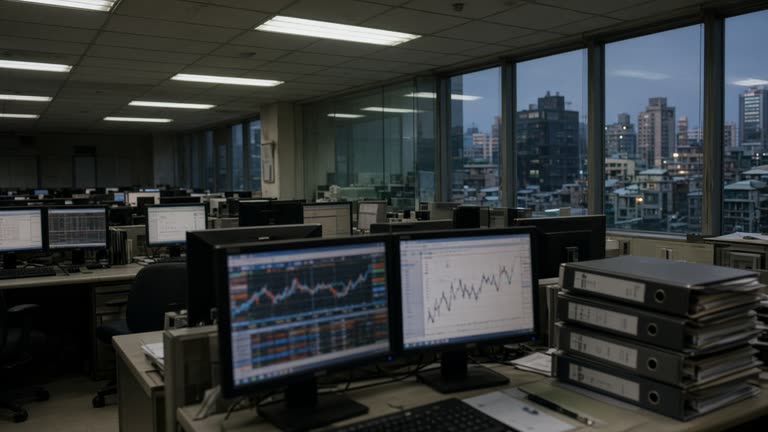 Taipei brokerage office with rows of desktop monitors showing blurred charts, fluorescent ceiling panels, glass partition and stacked compliance folders on a desk, evening light through windows, no logos or readable text