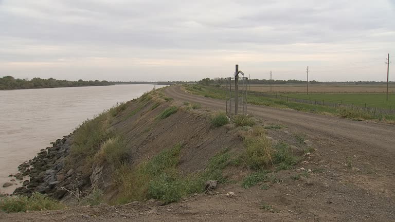 Murray River town levee with farmland and a fenced pipeline marker post in middle distance under hazy afternoon light, no logos or readable text
