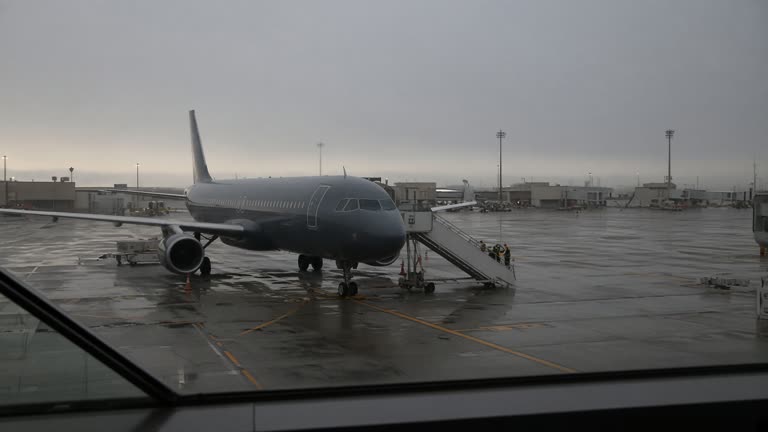 Dawn view through airport terminal glass of a narrow-body jet parked at a gate with mobile stairs, wet grey tarmac and soft overcast light, blurred ground vehicles, no readable airline logos.