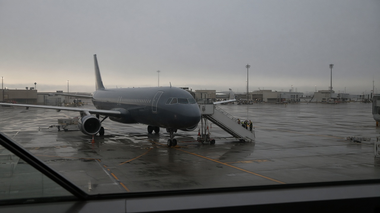 Dawn view through airport terminal glass of a narrow-body jet parked at a gate with mobile stairs, wet grey tarmac and soft overcast light, blurred ground vehicles, no readable airline logos.