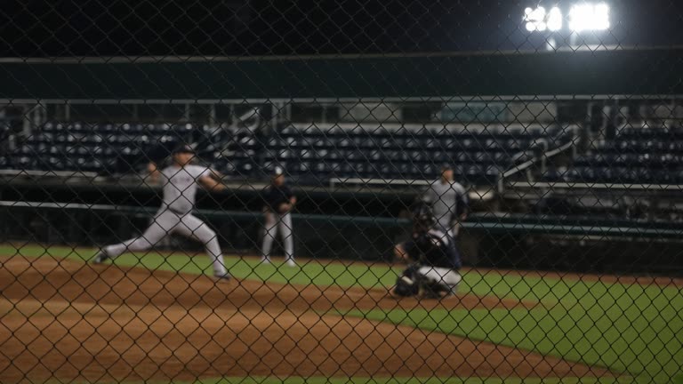 Baseball bullpen at night under stadium floodlights, blurred pitchers warming beyond chain-link fence, empty green seats and chalk foul line, no readable signage or team logos.
