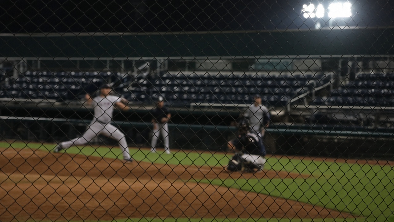 Baseball bullpen at night under stadium floodlights, blurred pitchers warming beyond chain-link fence, empty green seats and chalk foul line, no readable signage or team logos.