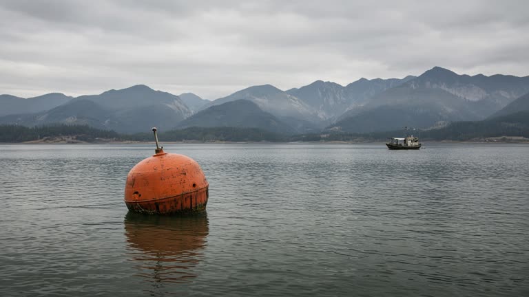 Orange research buoy on a calm freshwater lake with distant mountains and a small fishing boat, overcast morning, no logos or readable text.