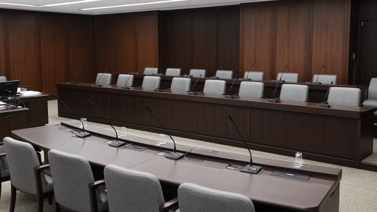 Japanese parliamentary committee hearing room with microphones on a long table and empty member seats, overhead fluorescent light, no logos or readable text.