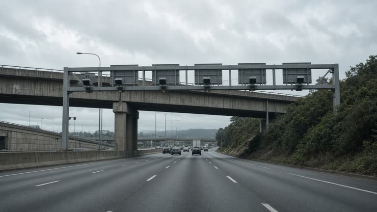 Sydney motorway toll gantry and concrete overpass in afternoon overcast light, muted traffic lanes, grey asphalt and green roadside scrub, no readable signage or logos