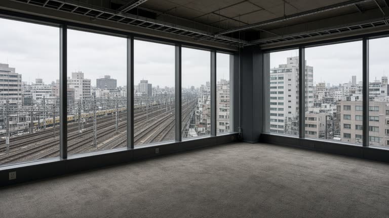 Empty corner office floor in a Tokyo high-rise with floor-to-ceiling windows overlooking rail tracks and overcast sky, bare carpet and cable trays, no logos or readable text