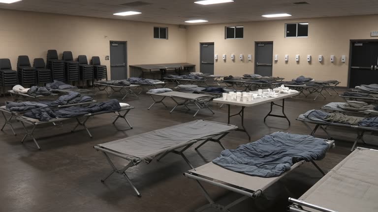 Municipal evacuation hall gymnasium with folding cots, wall-mounted hand sanitizer stations, and a small table holding sealed powder packets and paper cups, fluorescent lighting, no logos or readable text.