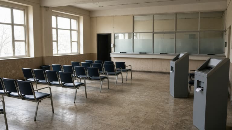 Public housing authority service hall with numbered ticket dispensers and waiting chairs facing a frosted glass counter, afternoon light through tall windows, worn linoleum floor, no logos or readable text