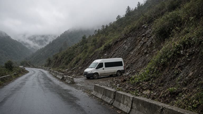 Rain-soaked mountain road with concrete debris barriers and disturbed hillside vegetation under low cloud, white survey vehicle parked on gravel pullout, no logos or readable text