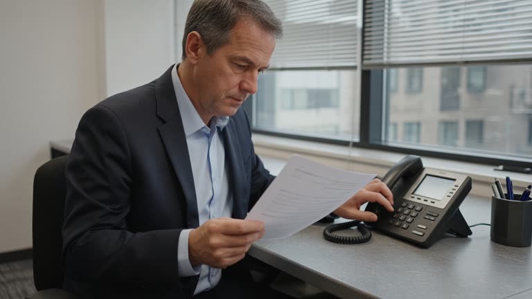 Corporate treasurer at a desk phone verifying a wire transfer printout beside a desktop banking terminal, gray office carpet, window light, no logos or readable text.