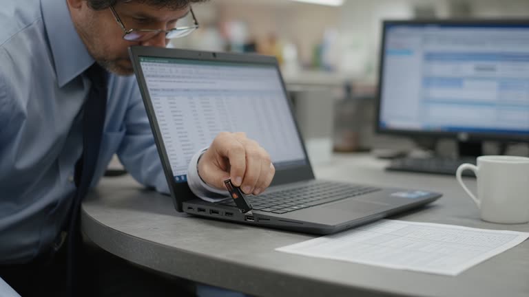 Accountant at a laminate desk inserting a USB security key into a laptop with a blurred second monitor, fluorescent office lighting, coffee mug and paper ledger, no logos or readable text