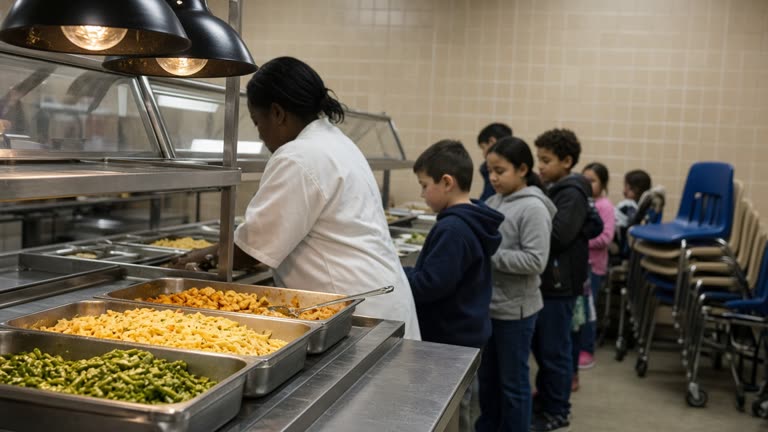 Elementary school cafeteria serving line with stainless trays and heat lamps over metal rails, children-sized chairs stacked against a tiled wall, flat fluorescent lighting, no logos or readable text