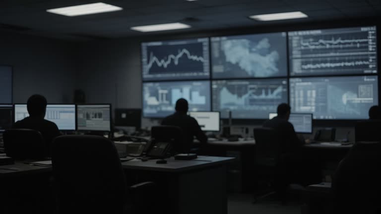 Dim operations room with wall-mounted status monitors showing blurred graphs, operator silhouettes at desks, cool monitor glow and overhead fluorescent mix, no readable text or logos