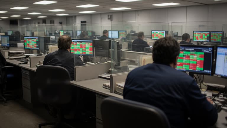 Retail brokerage office with rows of desktop monitors showing blurred green and red price tiles, evening fluorescent light, plastic partition dividers, no logos or readable text.