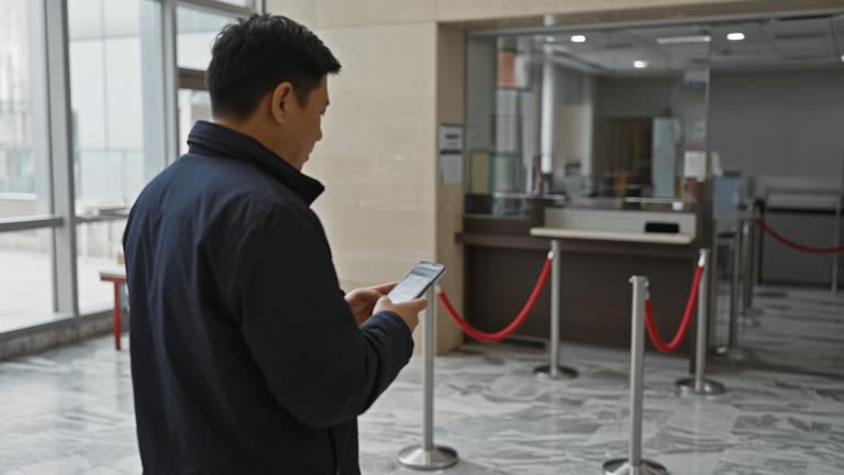 Consumer at a bank branch kiosk holding a smartphone with a blurred screen, marble floor and queue rope barriers under daylight, no logos or readable text