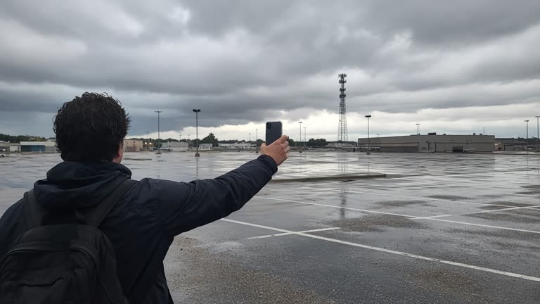 A person holding a smartphone at arm’s length in a rain-slick Gulf Coast parking lot, distant cell tower and low storm clouds, wet asphalt reflections, muted afternoon light, no logos or readable text