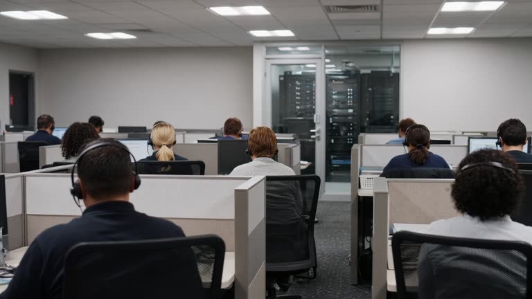 Government contact centre floor with agents at cubicles wearing headsets, server glass door visible in background, flat overhead lighting, faces turned away, no readable screens or logos