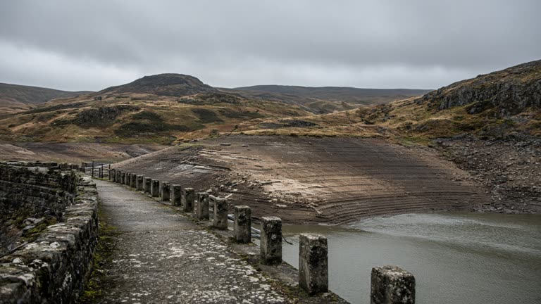 Low waterline on a stone-lined upland reservoir in Cumbria under grey September cloud, exposed shoreline and moor grass, maintenance walkway in foreground, no logos or readable text