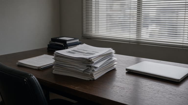 Law office conference table stacked with redacted paper briefs and a closed laptop, venetian blinds casting stripes, neutral walls, no logos or readable text