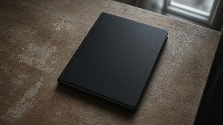 Closed unlabeled black hardcover notebook face-down on a wood desk, blank cover, overcast window light, no people, no logos, no readable text