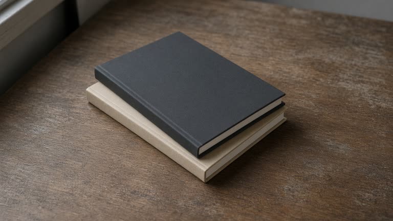Closed unlabeled black hardcover notebook face-down on a wood desk, blank cover, overcast window light, no people, no logos, no readable text