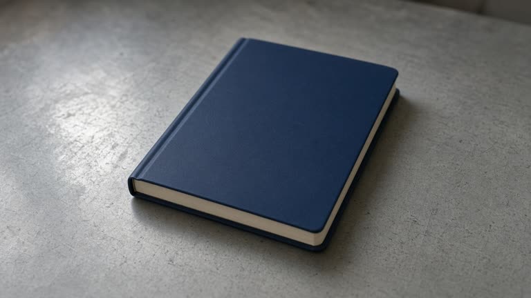 Closed unlabeled black hardcover notebook face-down on a wood desk, blank cover, overcast window light, no people, no logos, no readable text