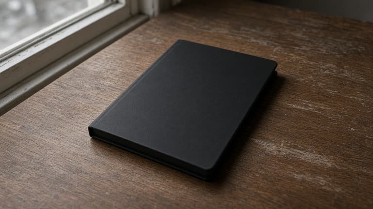 Closed unlabeled black hardcover notebook face-down on a wood desk, blank cover, overcast window light, no people, no logos, no readable text