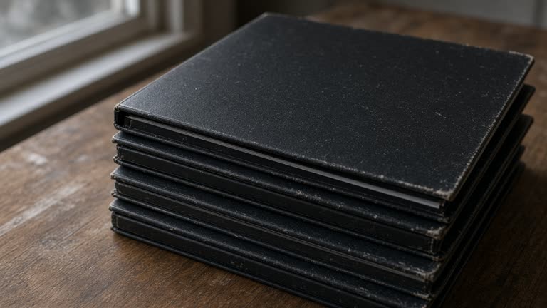 Close-up of closed blank black binders stacked on a wood table, overcast window light, no people, no logos, no readable text