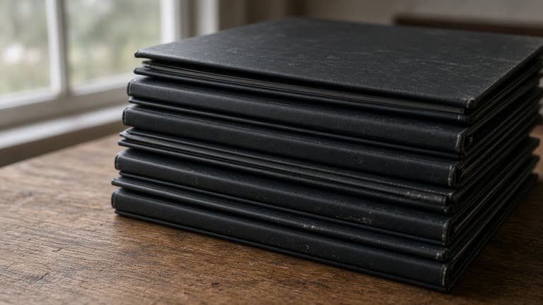 Close-up of closed blank black binders stacked on a wood table, overcast window light, no people, no logos, no readable text