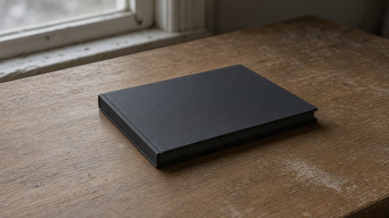 Closed unlabeled black hardcover notebook face-down on a wood desk, blank cover, overcast window light, no people, no logos, no readable text