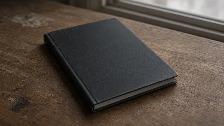 Closed unlabeled black hardcover notebook face-down on a wood desk, blank cover, overcast window light, no people, no logos, no readable text