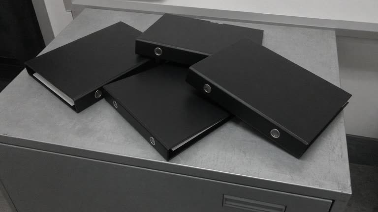 Close-up of closed blank black binders stacked on a wood table, overcast window light, no people, no logos, no readable text