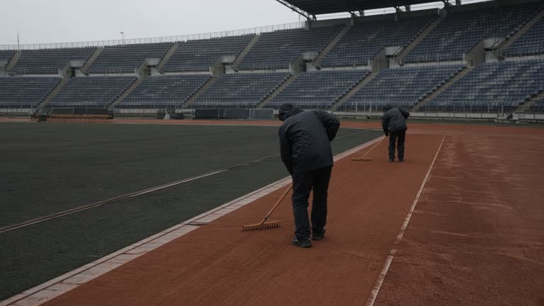 Empty athletics stadium javelin runway after light rain with ground crew raking clay surface, overcast sky, distant seating bowl without spectators, no logos or readable text