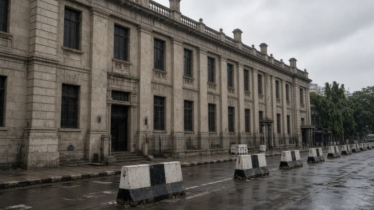 Reserve Bank of India headquarters stone facade on Mint Street in Mumbai under overcast monsoon sky, security barriers and wet pavement, no logos or readable text