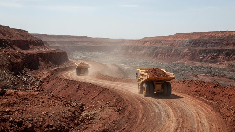 Wide view of red earth open-pit mine with yellow haul trucks on a haul road, harsh midday sun, dust haze, no logos or readable text
