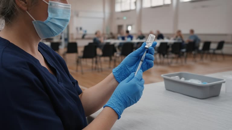 A community hall set up as a pop-up vaccination clinic, rows of plastic chairs on a timber floor, a nurse in blue gloves drawing a dose from a small glass vial into a syringe in the foreground, soft afternoon light through high windows, no logos or readable text