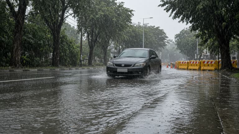 Afternoon rain on a tree-lined urban road in Singapore, ankle-deep water covering the curb lane, sedan crawling through, yellow roadwork barriers in background, no readable shop signs