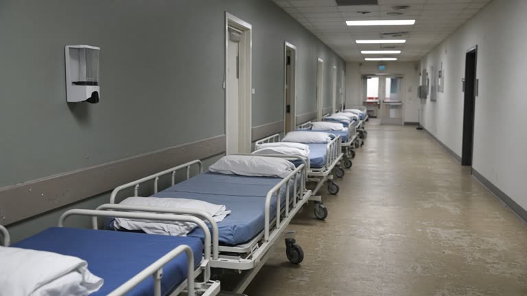 Pediatric hospital corridor with portable cots lined along one wall, a wall-mounted hand sanitizer dispenser, and soft overhead lighting on worn linoleum, no logos or readable text